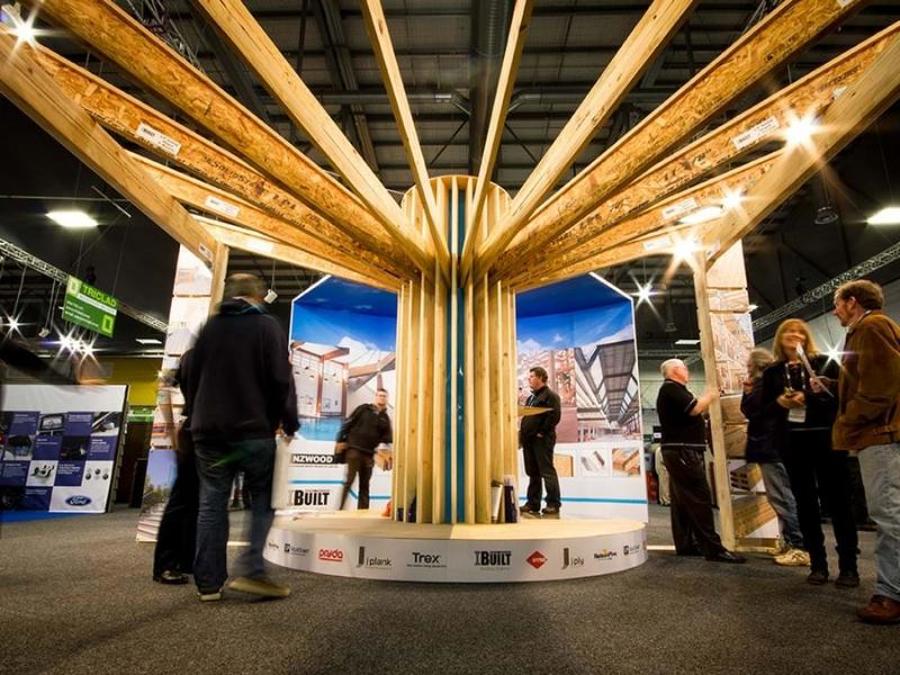 NZWood and iBuilt exhibition stand with timber beams radiating from a central timber column above a round platform, with product image panels and visitors walking around it
