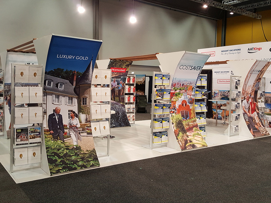 Travel brand exhibition stand on a white floor with curved fabric graphic towers and brochure racks, in an exhibition hall