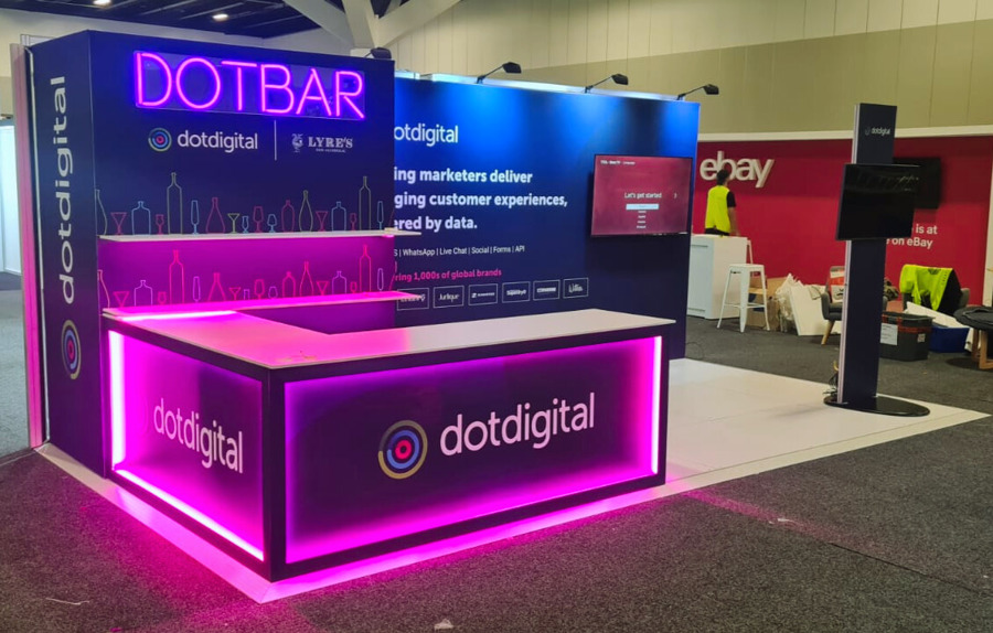 dotdigital exhibition stand with a magenta-lit bar counter, a neon DOTBAR sign, a wall-mounted screen and a screen on a freestanding post