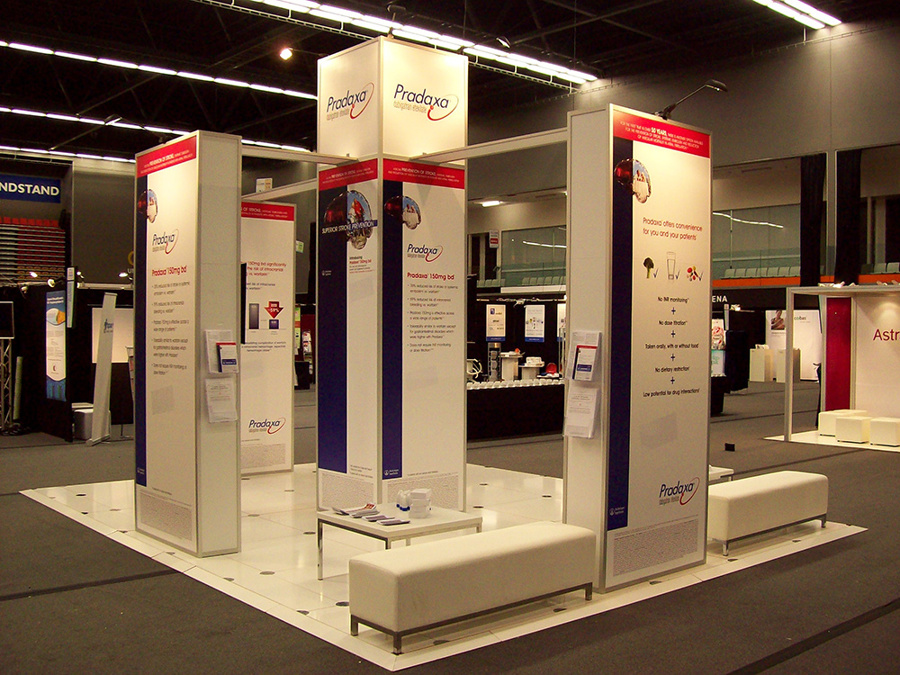 Island exhibition stand on a white raised floor with a visible edge, tall graphic columns and white bench seating, in an exhibition hall