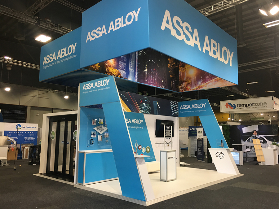 ASSA ABLOY exhibition stand built on a white raised floor, with a suspended blue hanging sign above
