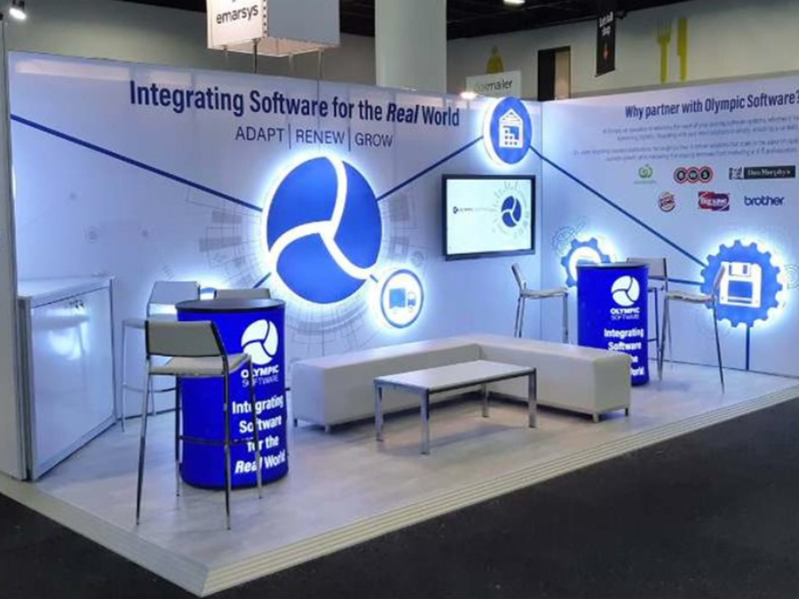 Corner exhibition stand with a white lounge seating area, high tables with stools, a wall screen and backlit circular logo graphics