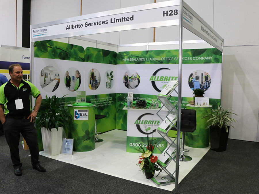 Corner shell scheme stand at a facilities management trade show with full-height printed wall graphics, a branded counter, round green plinths and a brochure stand