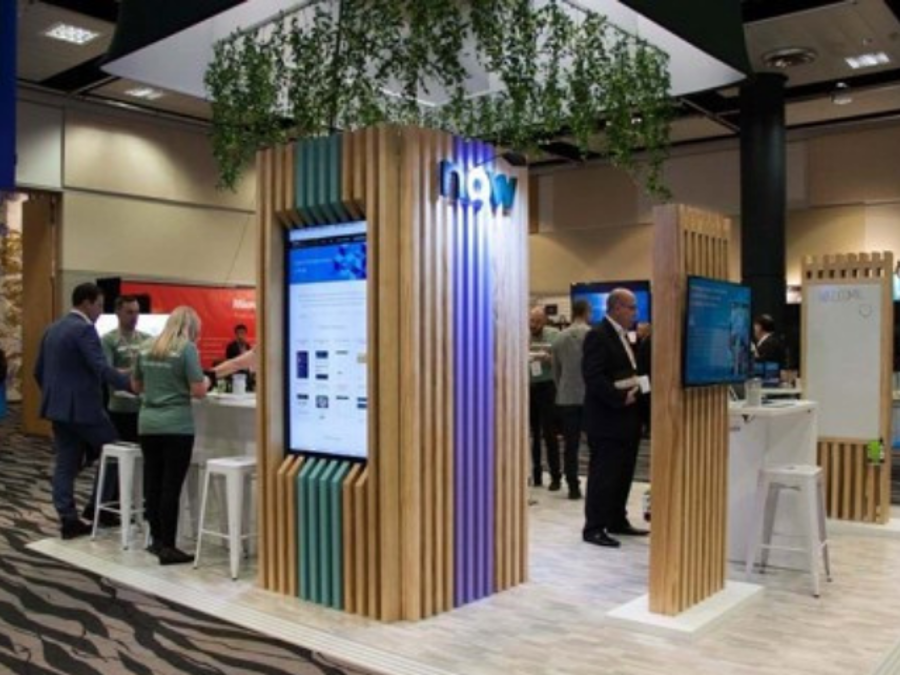 ServiceNow exhibition stand with timber-batten towers holding a portrait screen and a wide screen, and staff talking with visitors at white high tables