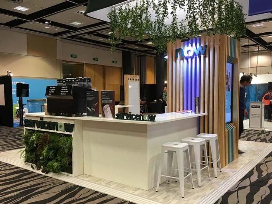 ServiceNow exhibition stand with a white coffee bar counter, white stools, a planter wall of ferns, a timber-slat column with the illuminated "now" logo and hanging greenery overhead
