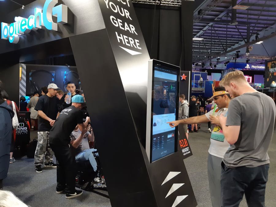Visitors using a tall freestanding touchscreen kiosk at a gaming expo, with a seated gaming rig in use behind them